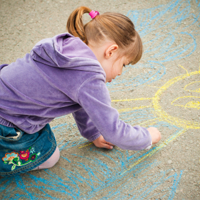 Ein kleines Mädchen mit einem lilafarbenen Pullover malt mit Straßenkreide Himmel und Sonne auf den Asphalt.